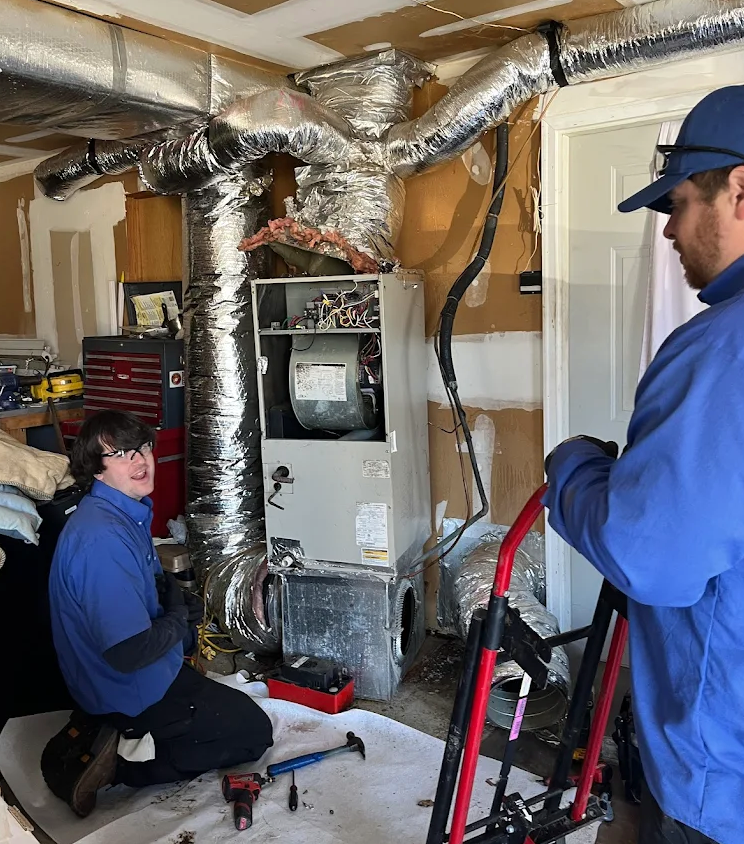 Two ARS HVAC technicians working on a furnace, with visible ductwork and tools on the floor.